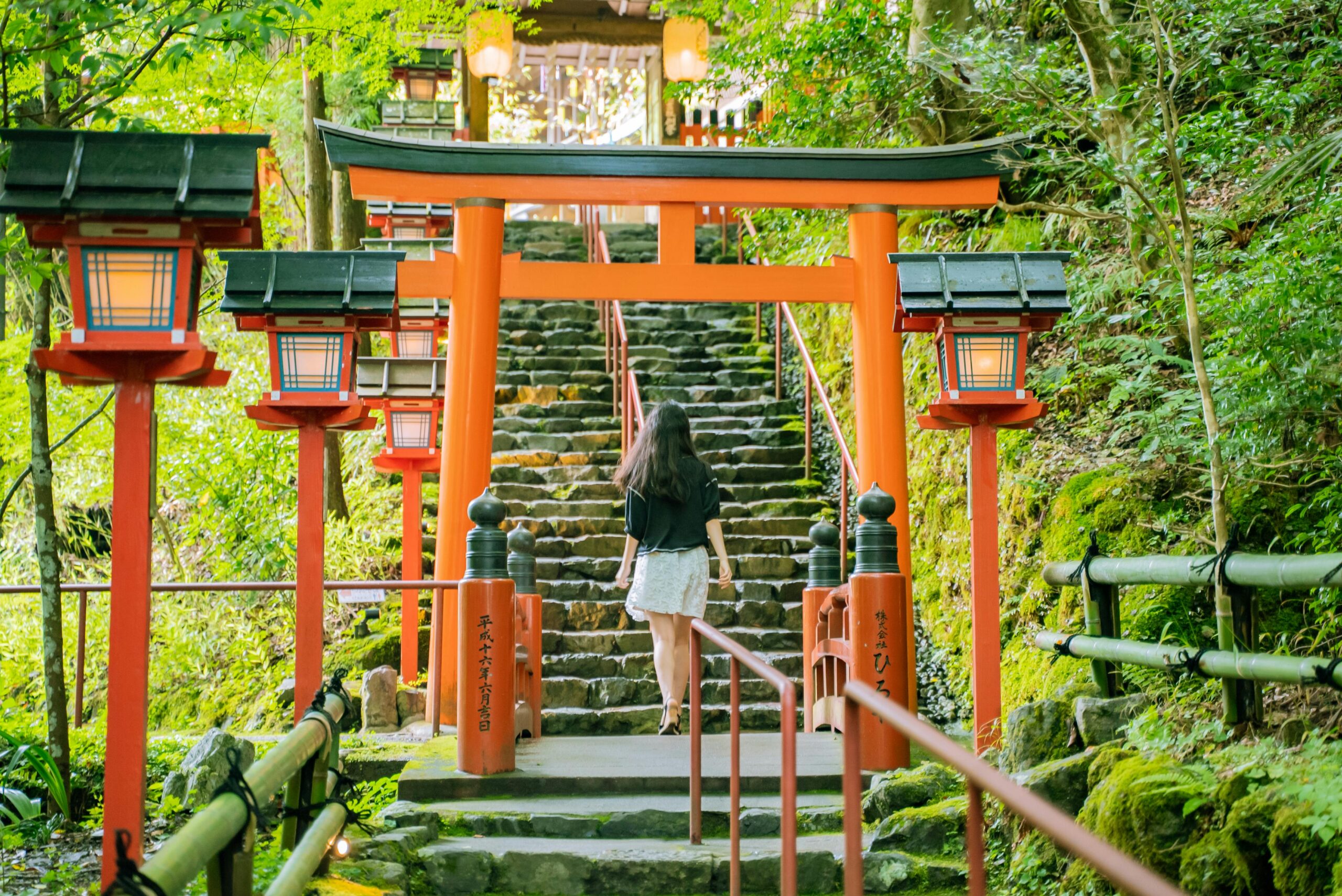 A woman walking up stone steps towards a traditional Japanese Shinto shrine surrounded by lush greenery.
